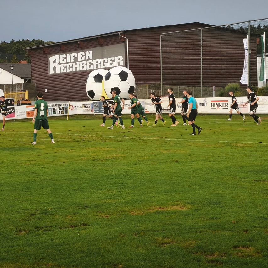 Ein Fußballspiel auf einem Feld mit Spielern in grünen und schwarzen Uniformen. Ein großer Fußball ist auf dem Gebäude hinter ihnen angebracht.