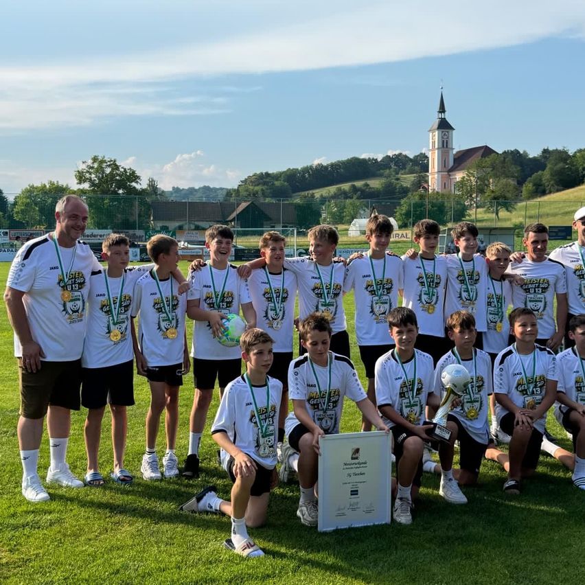 Eine Gruppe junger Fußballer posiert mit ihren Trainern, Medaillen und Trophäen auf einem Feld. Sie tragen weiße Shirts und Shorts. Ein Pokal und ein Zertifikat stehen vor ihnen. Dahinter sind eine Kirche und ein Zaun.