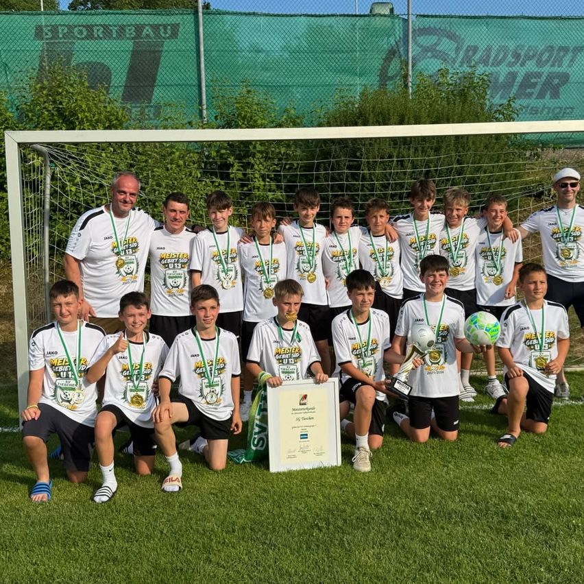 Eine Fußballmannschaft posiert für ein Gruppenfoto, sie tragen weiße Trikots mit Medaillen um den Hals. Ein Trainer steht hinter ihnen und eine Trophäe ist in der Mitte. Der Hintergrund zeigt ein Fußballtor und eine grüne Banner.