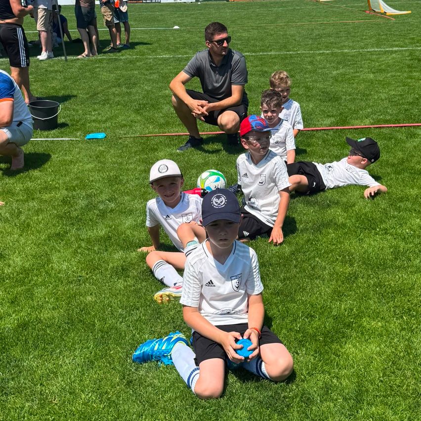Mehrere Jungen sitzen auf dem Rasenfeld. Ein Mann kniet hinter ihnen. In der Nähe der Jungen liegen ein paar Fußballbälle. Ein Eimer steht neben dem Mann. Einige Leute stehen in der Ferne.