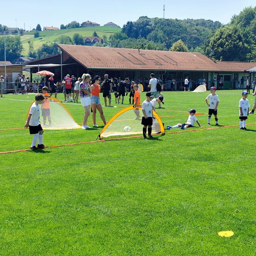 Kinder trainieren Fußball auf einem Feld. Eine Frau steht neben einem Fußballtor. Viele Menschen versammeln sich, um die Aktivität zu beobachten. Bäume und Häuser sind im Hintergrund.