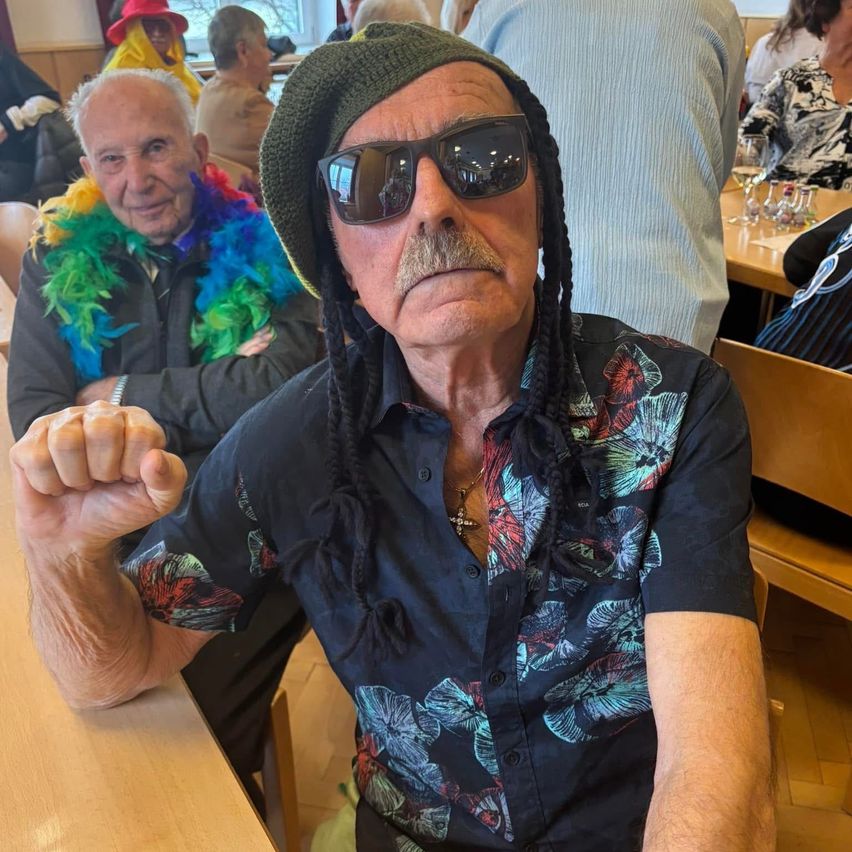 Ein älterer Mann mit buntem Hut und Sonnenbrille sitzt an einem Tisch und macht eine Faust. Hinter ihm lächelt ein anderer Mann mit einem Regenbogenschal. Die Szene spielt sich in einem Essbereich mit Holztischen und Stühlen ab.