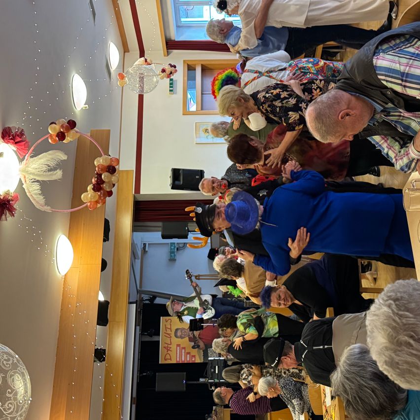 Eine Menschenmenge tanzt in einem Raum mit farbenfrohen Dekorationen und Luftballons, die von der Decke hängen. Eine Person spielt Gitarre auf der Bühne.