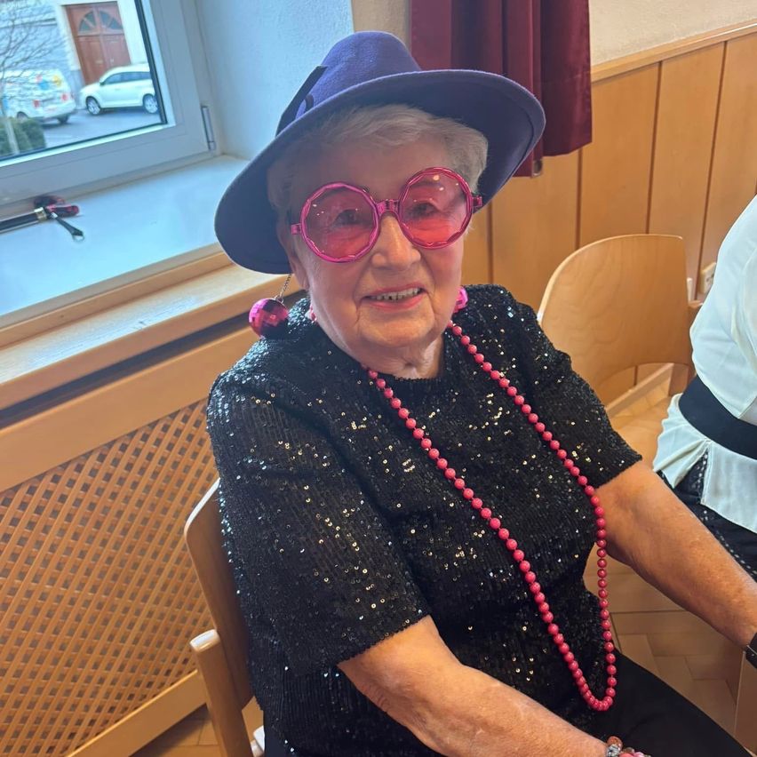 Eine ältere Frau mit einem lila Hut und rosa Sonnenbrille sitzt in einem Stuhl in einem Raum mit einer Holzwand und einem Fenster, das ein Auto draußen zeigt.