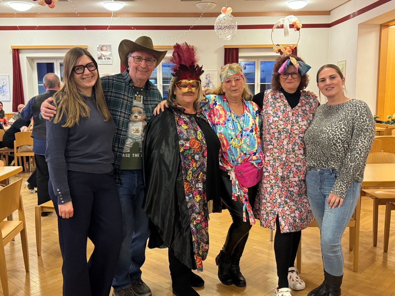 Eine Gruppe von Menschen in verschiedenen Kostümen steht zusammen und lächelt für ein Foto in einem Raum mit Holzböden und dekorativen Lichtern.