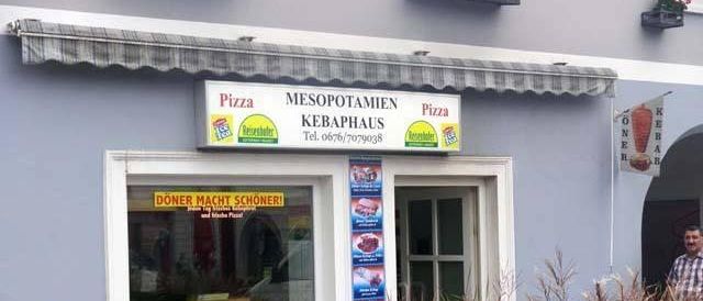 Ein Pizzaladen mit einem Schild Mesopotamien Kebaphaus, mit einem Cartoon-Charakter auf der linken Seite, einem stehenden Mann und Tischen und Stühlen draußen.