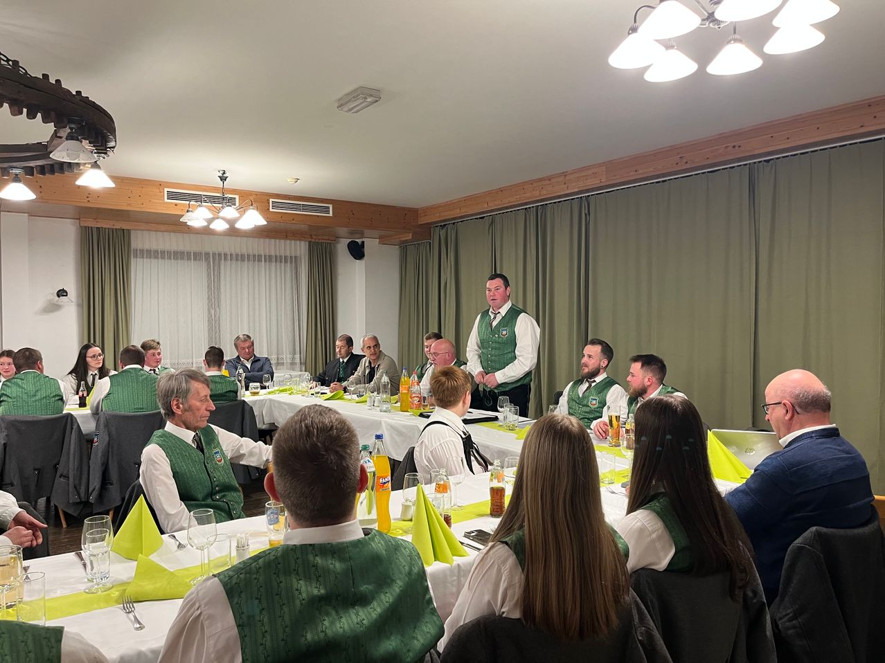 A group of people in green vests sit around a table in a meeting room. A man in a white shirt stands near a table with bottles and glasses.