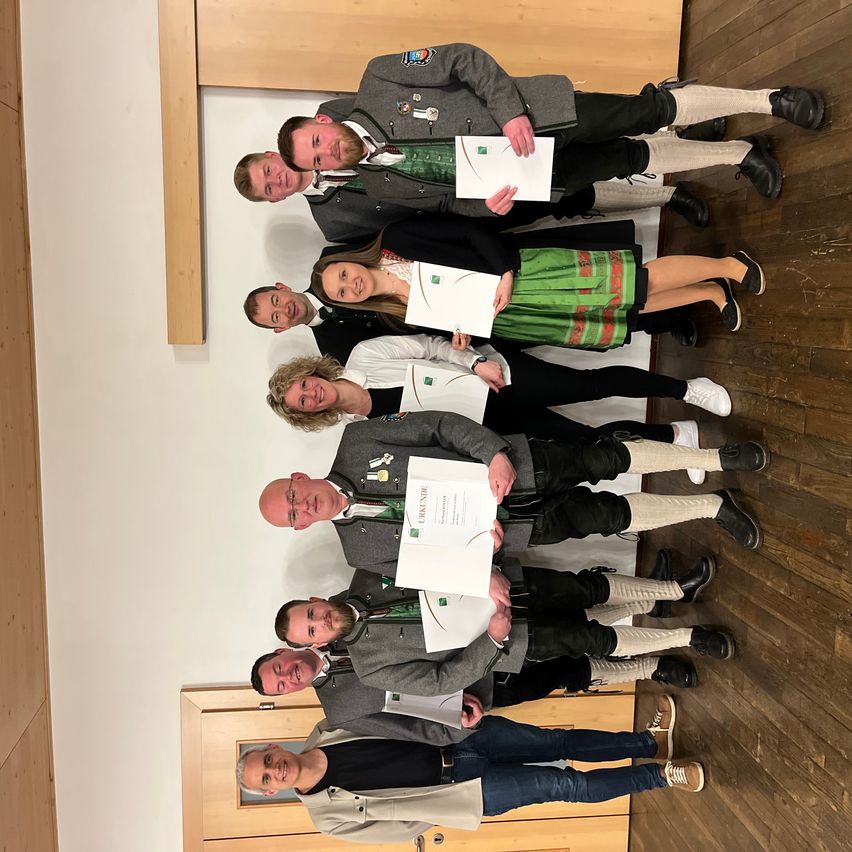 Seven people pose for a photo while holding certificates in their hands. They all seem to be smiling. They are wearing traditional attire. The room is wooden, and there are doors on the left and right.