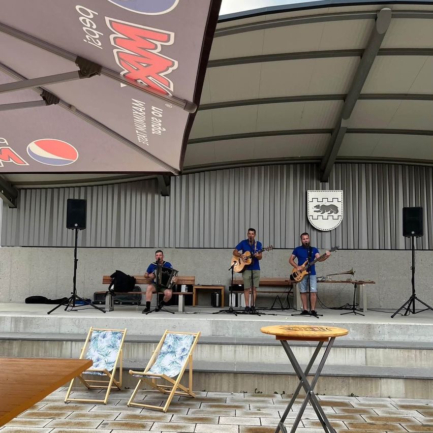 Drei Männer spielen auf einer Bühne mit Musikinstrumenten. Einer spielt Gitarre, ein anderer Akkordeon und der dritte Trompete. Es gibt Lautsprecher und eine Pepsi-Werbung auf der Bühne.