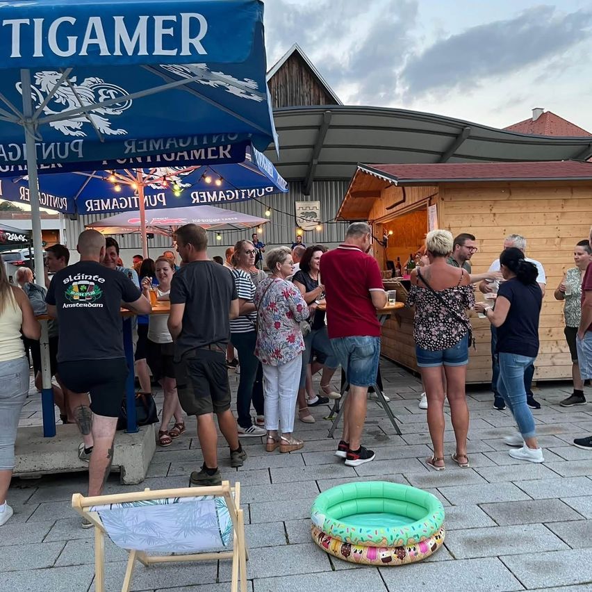 Eine Gruppe von Menschen versammelt sich vor einem Lokal mit blauen Schirmen und einem Schild, das 'TIGAMMER' besagt. Sie stehen und unterhalten sich um Tische herum, einige halten Getränke. Ein kleiner aufblasbarer Pool liegt auf dem Boden.
