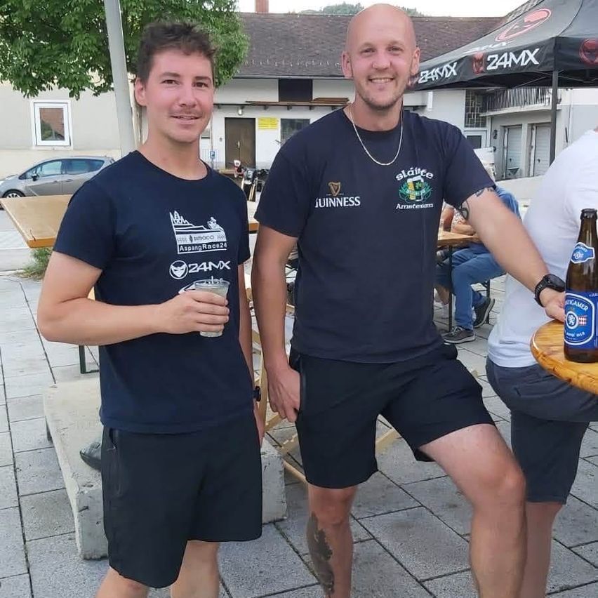 Zwei Männer in identischen blauen T-Shirts und schwarzen Shorts stehen draußen, einer hält ein Getränk und lächelt in die Kamera. Hinter ihnen ist ein Gebäude mit einem Zelt und einem Auto zu sehen.