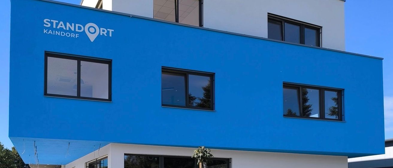 Ein modernes Gebäude mit blauer Fassade, 'STANDORT KAINDORF' in weißen Buchstaben, Glasfenstern und einem Weg zum Eingang.