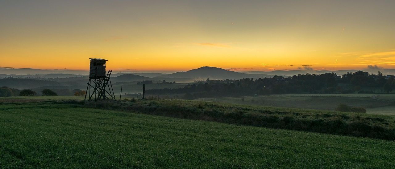 Eine ruhige ländliche Szene mit einem Jagdturm, der sich gegen einen warmen Sonnenuntergang abzeichnet. Hügel und eine klare, weitläufige Wiese unter einem blauen Himmel.