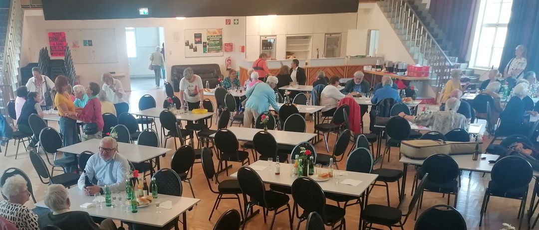 Ein Indoor-Restaurant mit mehreren Tischen und Stühlen, die von Menschen besetzt sind. Einige stehen in der Nähe der Tische. Die Tische haben Blumen, Flaschen und Essen. Der Raum hat eine Treppe und Schränke. Es gibt einen Feuerlöscher an der Wand befestigt.