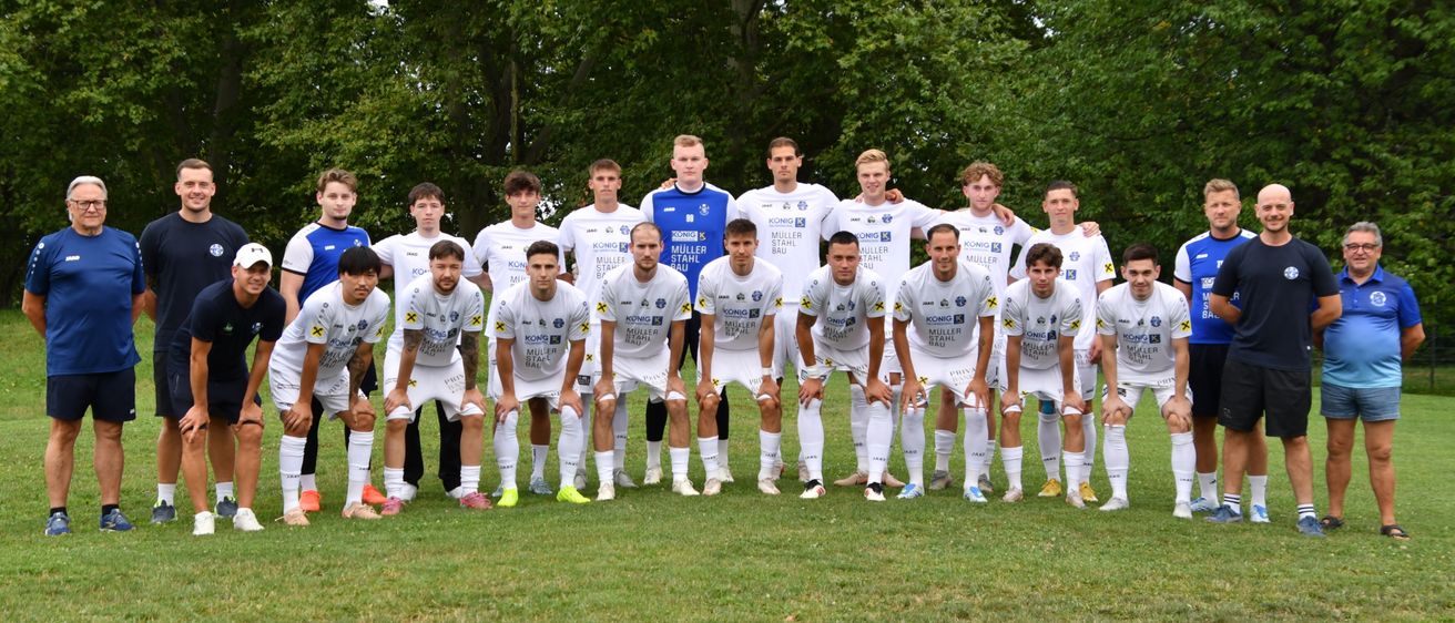 Eine Fußballmannschaft in weißen Trikots mit gelben Sternen auf der Vorderseite posiert für ein Foto auf einem Rasenfeld mit Bäumen im Hintergrund.