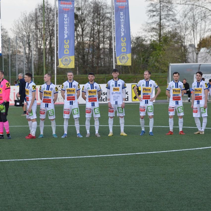 Neun Fußballspieler in weißen und blauen Trikots mit den Buchstaben Rex auf ihren Trikots stehen auf einem Feld. Hinter ihnen befinden sich zwei Banner mit dem Wort DSC.