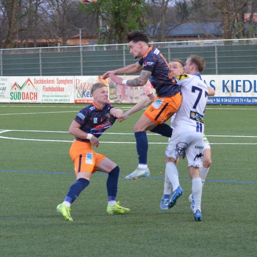Zwei Fußballspieler kollidieren, während einer versucht, den Ball zu köpfen. Ein Spieler trägt Blau und Orange, der andere trägt Weiß.