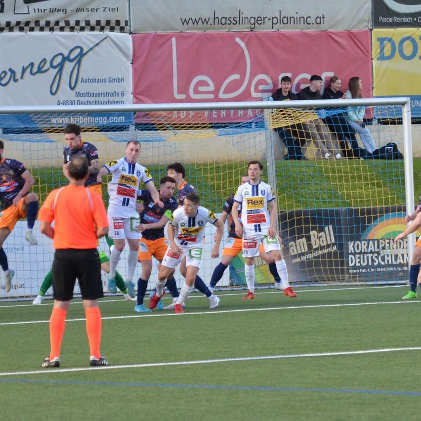 Fußballspieler in verschiedenen Trikots spielen auf einem Feld. Ein Spieler in einem orangefarbenen Trikot tritt den Ball. Ein Mann in einem orangefarbenen Shirt steht in der Nähe und beobachtet das Spiel.