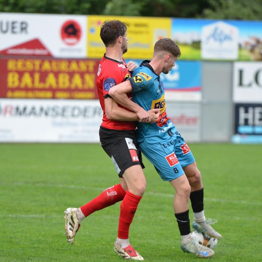 Zwei Fußballspieler konkurrieren auf einem Feld, einer trägt ein rotes Trikot und der andere ein blaues. Sie zielen beide auf den Ball in der Nähe ihrer Füße.