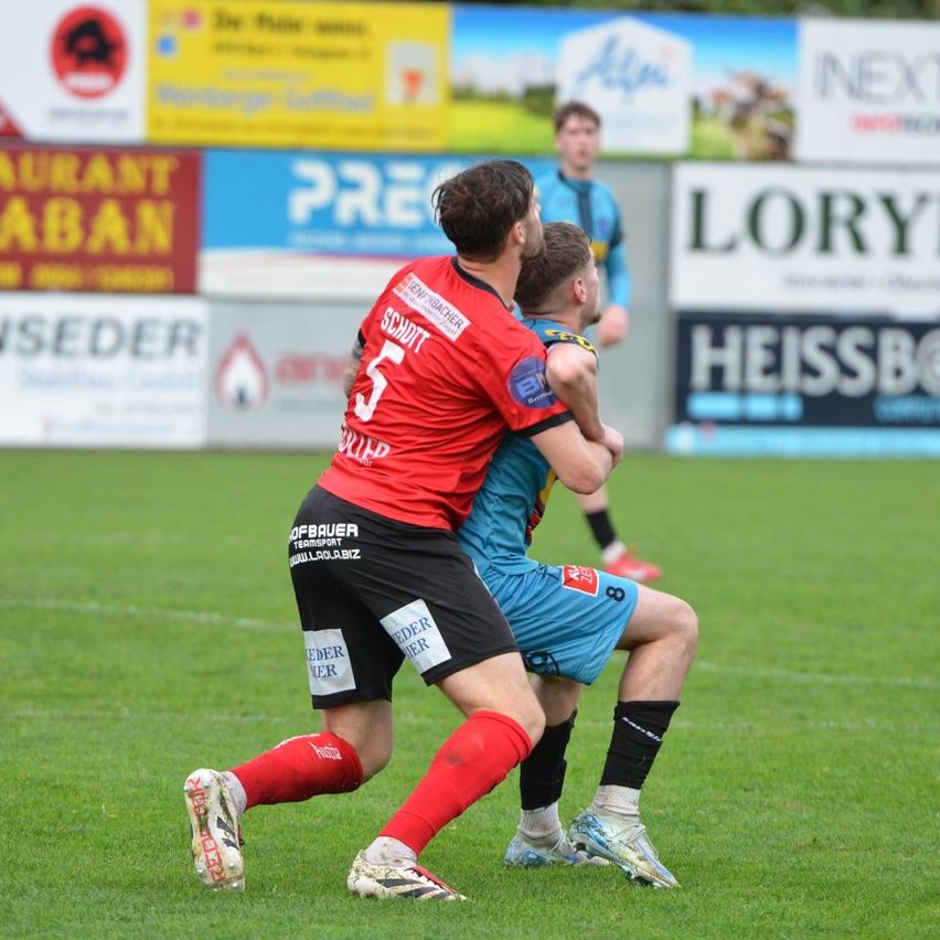 Zwei Fußballspieler spielen auf einem Feld. Einer trägt ein rotes Trikot mit der Nummer 5, der andere ein blaues Trikot mit der Nummer 8. Sie sind in einen körperlichen Wettkampf verwickelt.