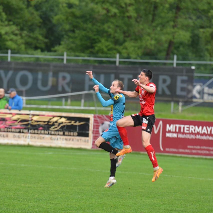 Zwei Fußballspieler springen während eines Spiels in der Luft, einer in Blau und einer in Rot. Hinter ihnen ist ein Zaun mit einem Banner zu sehen. Im Hintergrund befinden sich Bäume und ein Mann in einem blauen Jackett.