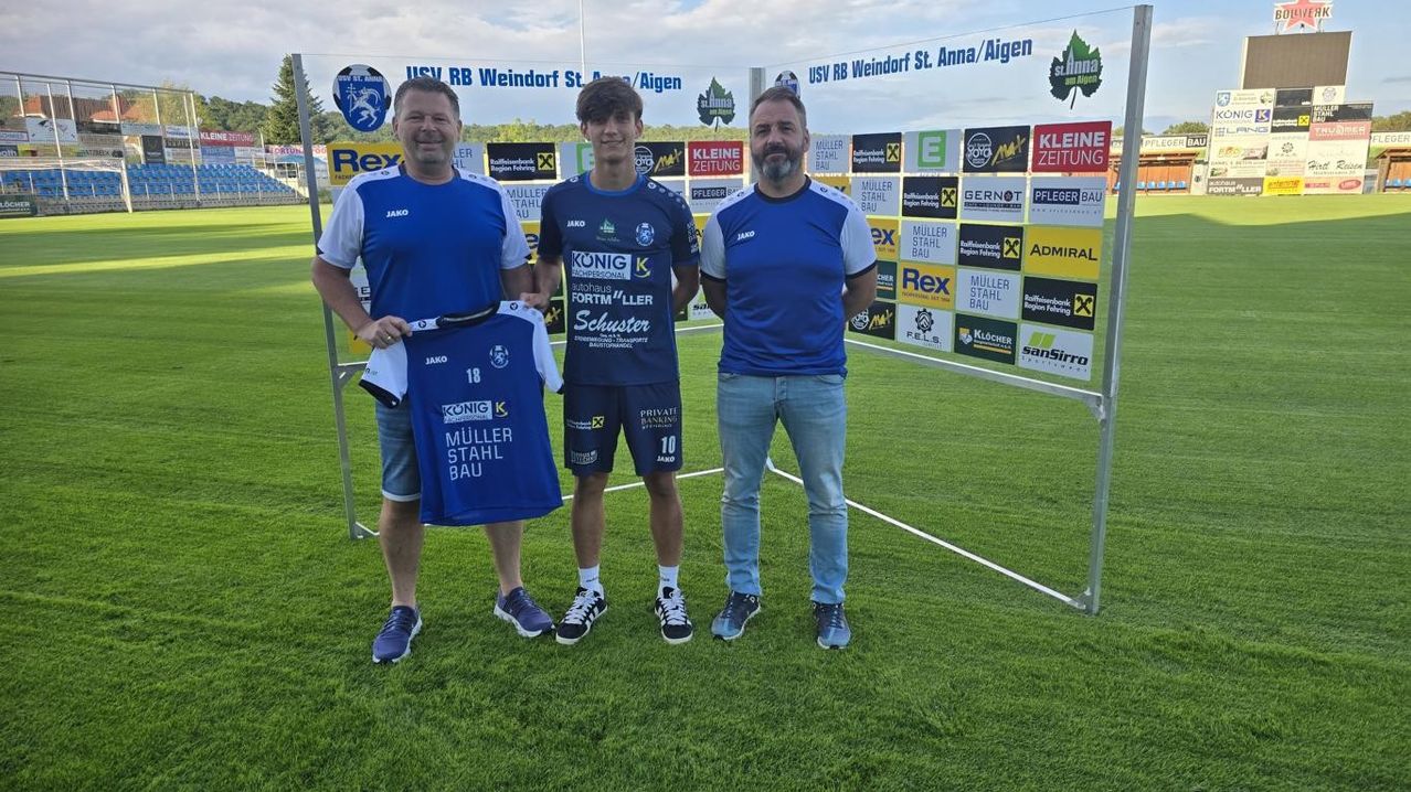 Drei Männer stehen auf einem Fußballfeld mit einer Werbekulisse. Der Mann auf der linken Seite hält ein blaues Trikot. Der Mann in der Mitte trägt ein blaues Trikot mit der Nummer 10. Der Mann auf der rechten Seite trägt ein blaues Hemd.