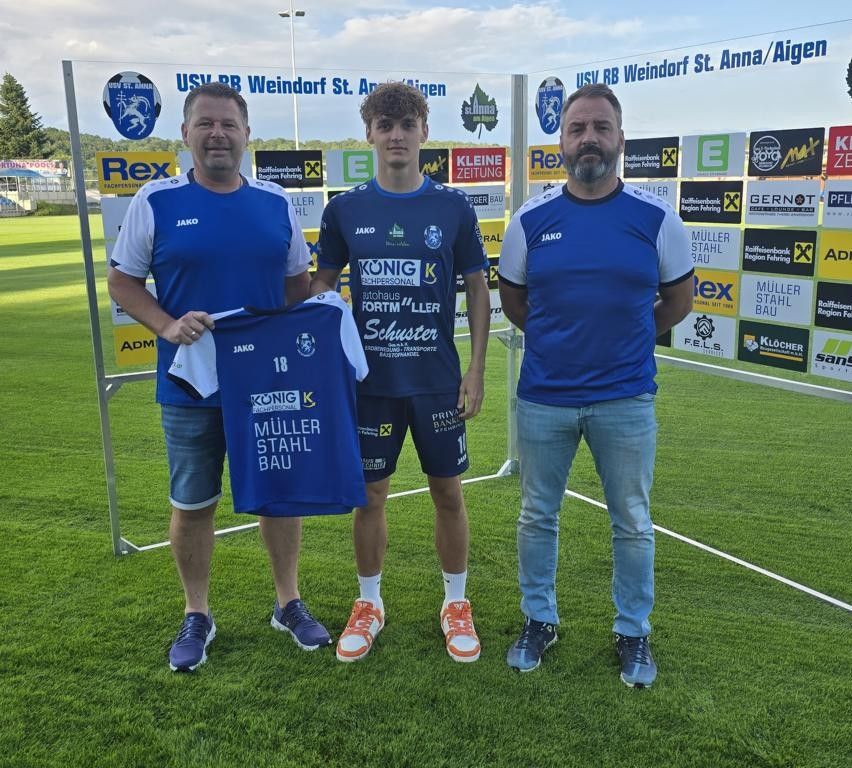 Drei Männer stehen auf einem Fußballfeld. Der Mann in der Mitte hält ein blaues Trikot mit der Nummer 18. Die anderen beiden Männer tragen blaue Trikots mit weißem Rand. Sie stehen vor einem Schild mit vielen Logos.