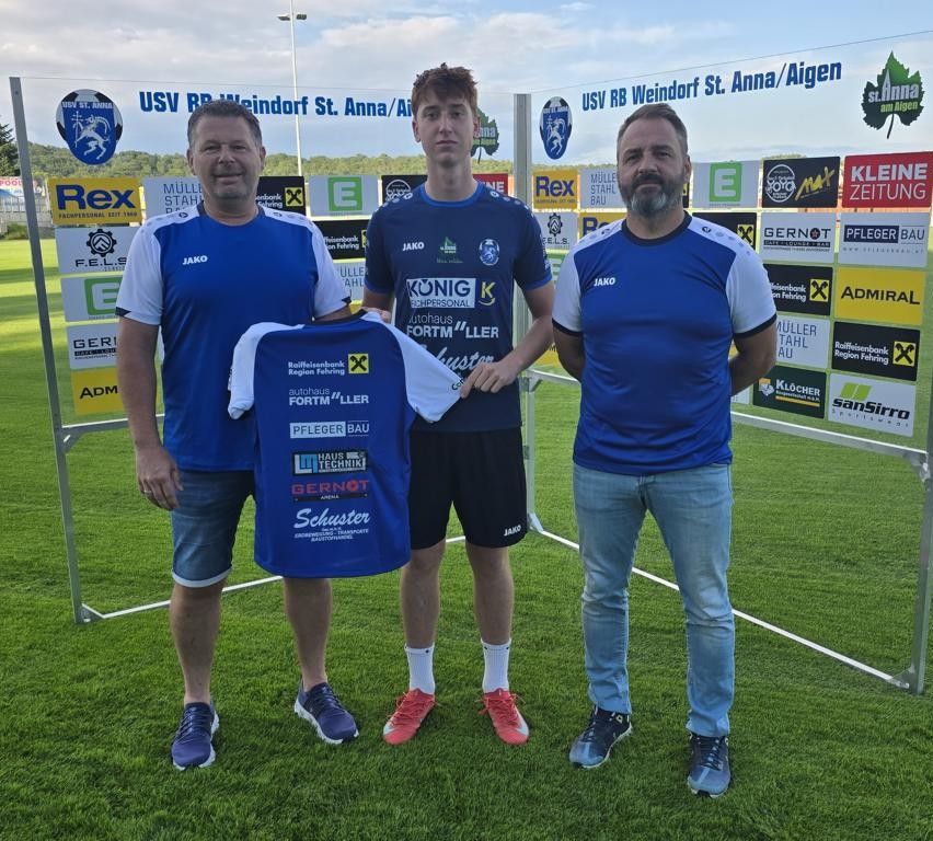 Drei Männer stehen auf einem Fußballfeld und tragen blaue Trikots mit weißen Säumen. Der Mann in der Mitte hält ein blaues Shirt mit Logos und Text. Hinter ihnen steht ein weißes Netz und ein Banner mit Werbung.