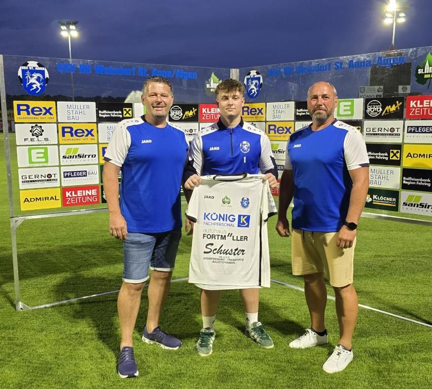 Drei Männer stehen bei Nacht auf einem Fußballfeld und lächeln für ein Foto. Der Mann in der Mitte hält ein weißes Trikot mit blauen Zahlen und Text. Die Männer tragen blaue Trikots, Shorts und Turnschuhe. Hinter ihnen ist eine Wand mit verschiedenen Logos und Texten.