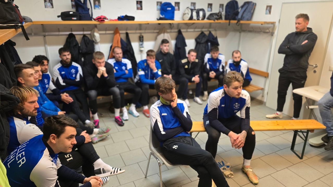 Eine Gruppe von Athleten in blau-weißen Uniformen sitzt auf Bänken in einer Umkleidekabine. Einige haben die Hände im Gesicht, andere schauen nach unten.