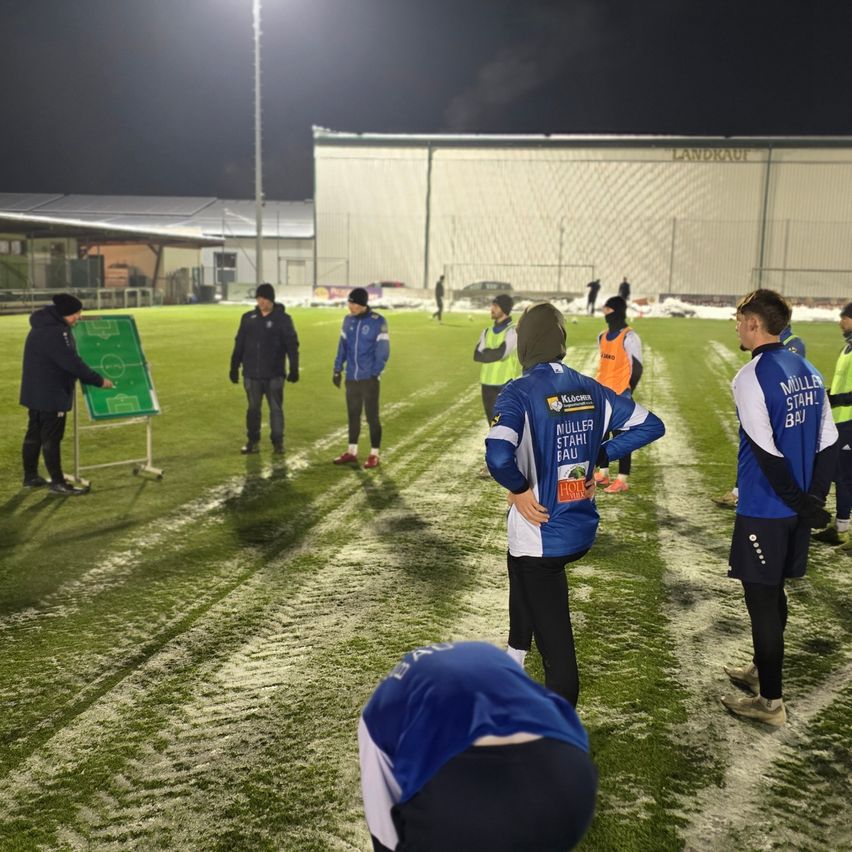 In der Nacht stehen mehrere Menschen in Winterkleidung auf einem Fußballfeld. Sie haben sich um einen Mann versammelt, der auf eine grüne Tafel zeigt.
