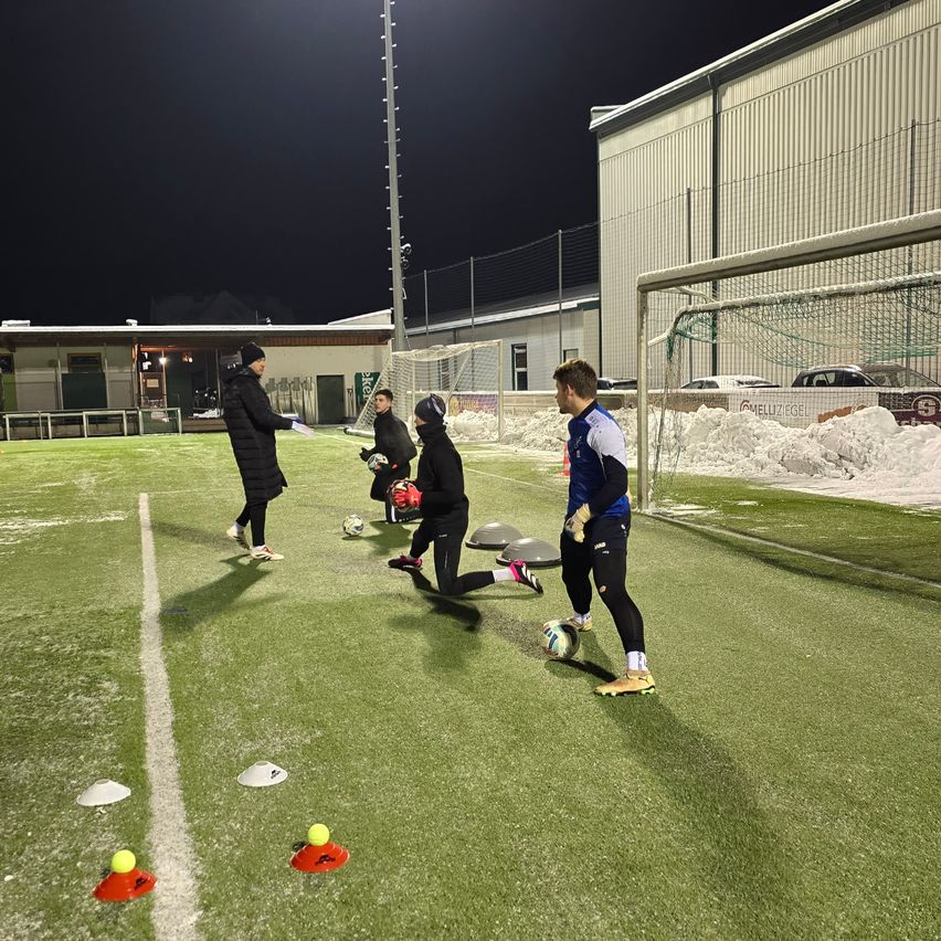 Mehrere Personen üben Fußball auf einem Feld bei Nacht. Sie tragen Winterkleidung. Es gibt Kegel und Bälle auf dem Feld. Das Feld hat eine weiße Linie und ein Tor.