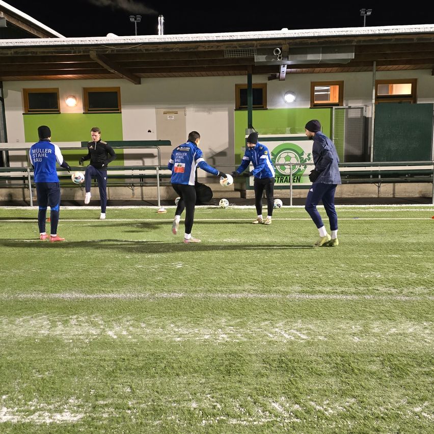 Eine Gruppe von Fußballspielern trainiert auf einem Feld mit grünem Rasen. Sie tragen blaue Uniformen. Das Feld hat weiße Markierungen. Dahinter steht ein Gebäude mit einer grünen Wand.
