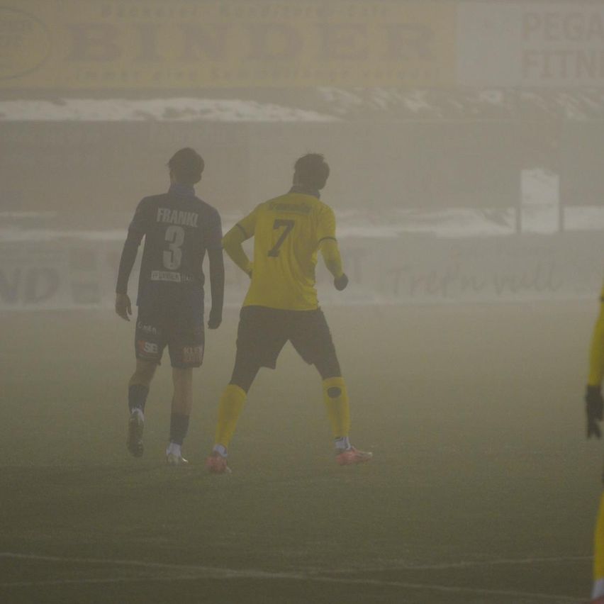 Zwei Fußballspieler bei nebligen Bedingungen, einer mit Trikotnummer 3 und der andere mit Nummer 7, laufen auf dem Feld.
