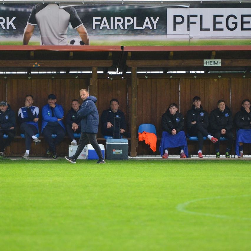 Eine Fußballmannschaft in blauen Uniformen sitzt auf einer Bank. Ein Mann in einer Kapuzenjacke geht vor ihnen. Ein Banner mit dem Text 'Fairplay Pflege' ist an der Wand.