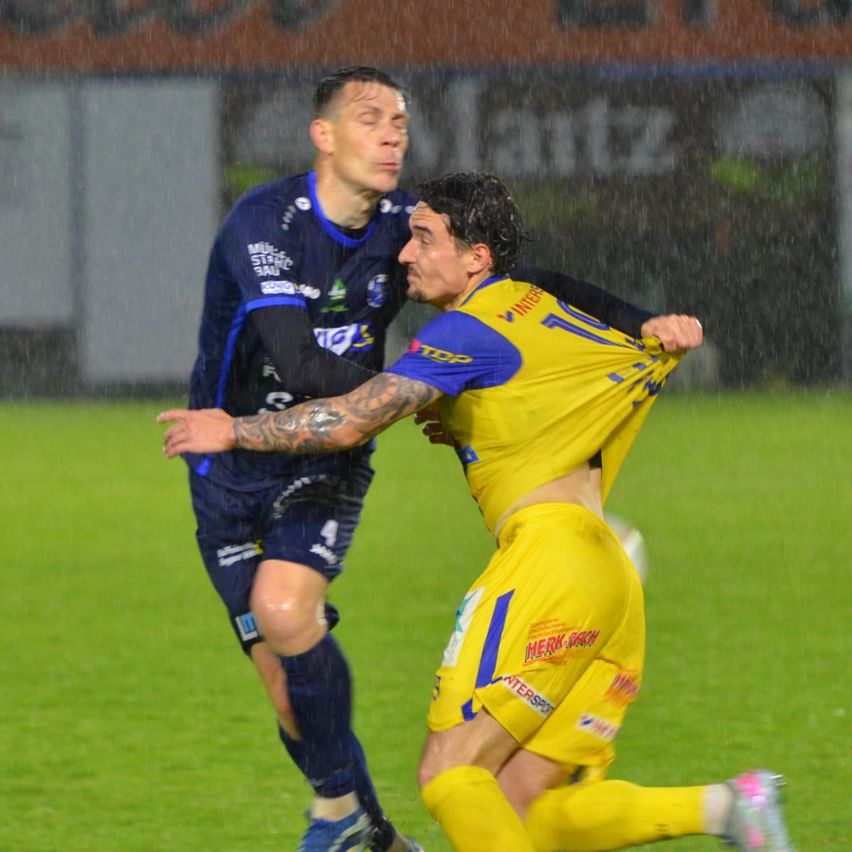 Zwei Fußballspieler in blauen und gelben Trikots sind auf einem Feld in eine körperliche Auseinandersetzung verwickelt, während eines Regenspiels.
