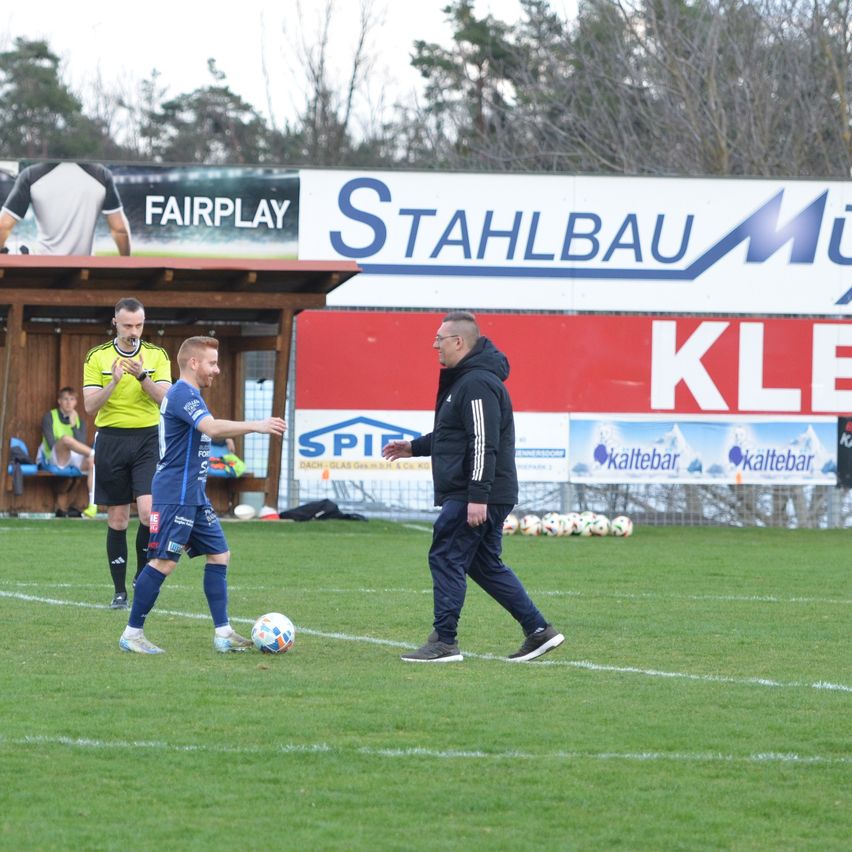 Ein Fußballspieler passt einen Ball auf dem Feld, während ein Trainer zusieht. Ein Schiedsrichter ist in der Nähe, und Werbung ist im Hintergrund sichtbar.