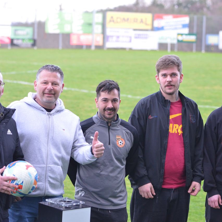 Eine Gruppe von Männern steht auf einem Rasenfeld, einer hält einen Fußball. Hinter ihnen sind mehrere Werbetafeln und ein Zaun. Sie alle lächeln und scheinen für ein Foto zu posieren.