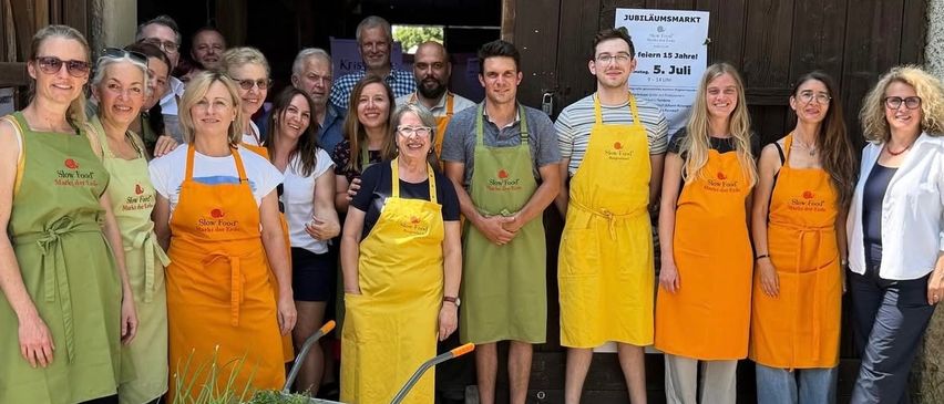 Eine Gruppe von Menschen in gelben Schürzen posiert für ein Foto vor einer Holztür. Einige halten eine Schubkarre mit Pflanzen. Ein Schild steht 'Jubilaumsmarkt'.