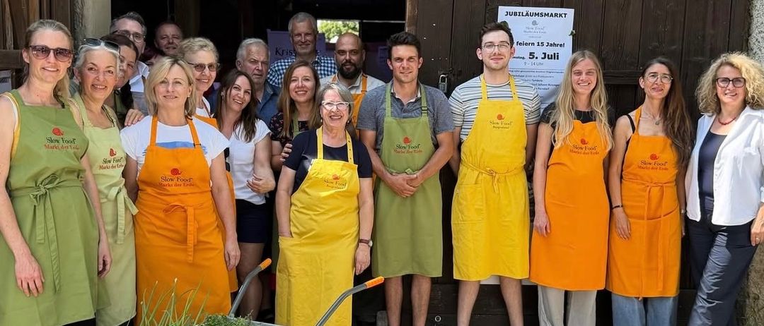 Eine Gruppe von Menschen in gelben Schürzen posiert für ein Foto vor einer Holztür. Einige halten eine Schubkarre mit Pflanzen. Ein Schild steht 'Jubilaumsmarkt'.