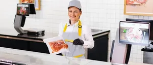 Eine Frau steht hinter einer Fleischtheke und lächelt. Sie hält ein verpacktes Fleischprodukt und ist in einem weißen Outfit und einer Schürze gekleidet. Verschiedene Fleischsorten sind auf der Theke ausgestellt.