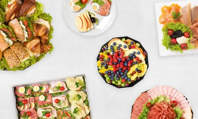Ein Tisch ist mit verschiedenen Gerichten geschmückt, darunter Sandwiches, Salate und eine Obstplatte. Im Mittelpunkt steht eine Obstsalat mit Blaubeeren, Trauben und Kirschen.