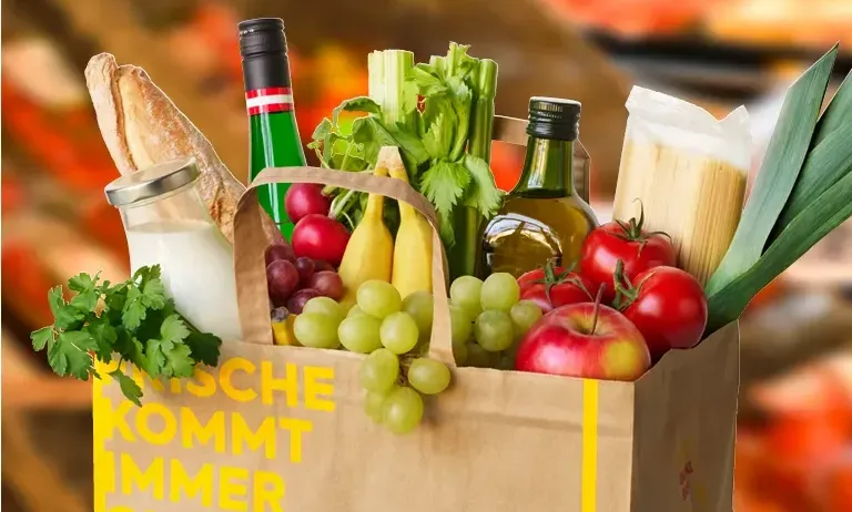 Ein brauner Papiersack mit verschiedenen Lebensmitteln im Inneren, darunter eine Flasche Wein, Brot und Gemüse. Der Sack ist mit den Worten 'Frische Kommt Immer Gut An' beschriftet.