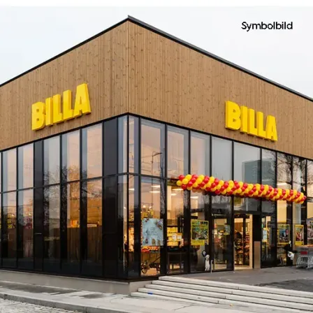 Die Vorderseite eines BILLA-Geschäfts mit großen gelben BILLA-Schildern, Glasfenstern und einem rot-gelben Luftballonbogen über dem Eingang.