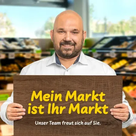 Ein Mann steht in einem Geschäft und hält ein hölzerne Schild mit gelbem Text, auf dem steht 'Mein Markt ist Ihr Markt. Unser Team freut sich auf Sie.' Er lächelt und scheint glücklich zu sein.