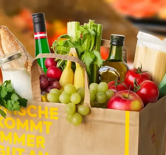 Ein brauner Papierbeutel ist mit verschiedenen Lebensmitteln gefüllt, darunter Brot, Wein, Trauben, Tomaten und Gemüse, und steht auf einem unscharfen Hintergrund. Der Text 'FRISCHE KOMMT IMMER GUT AN' ist auf dem Beutel gedruckt.