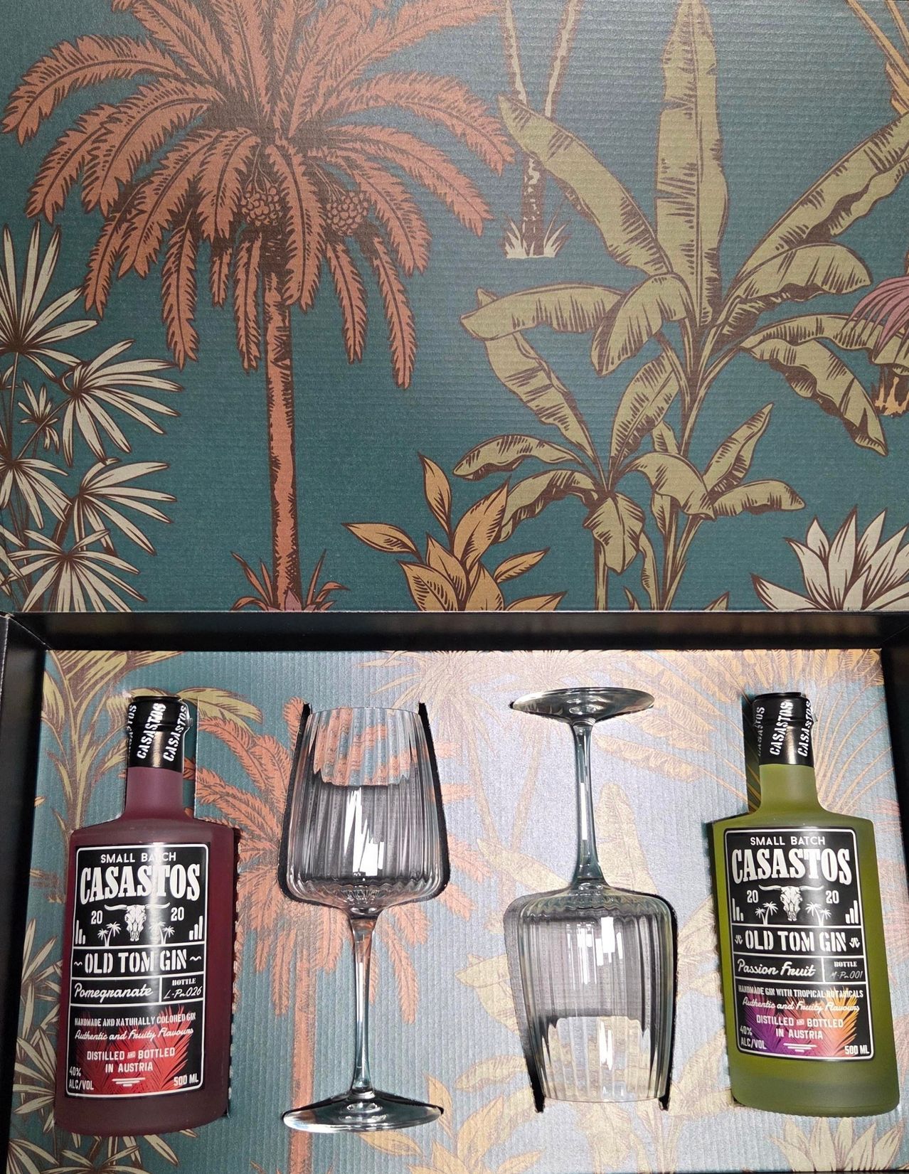 Zwei Flaschen Old Tom Gin und zwei Weingläser stehen auf einem Regal. Die Flaschen sind mit 'Small Batch' und 'Passion Fruit' beschriftet. Der Hintergrund hat ein tropisches Thema mit Palmen und Bananen.