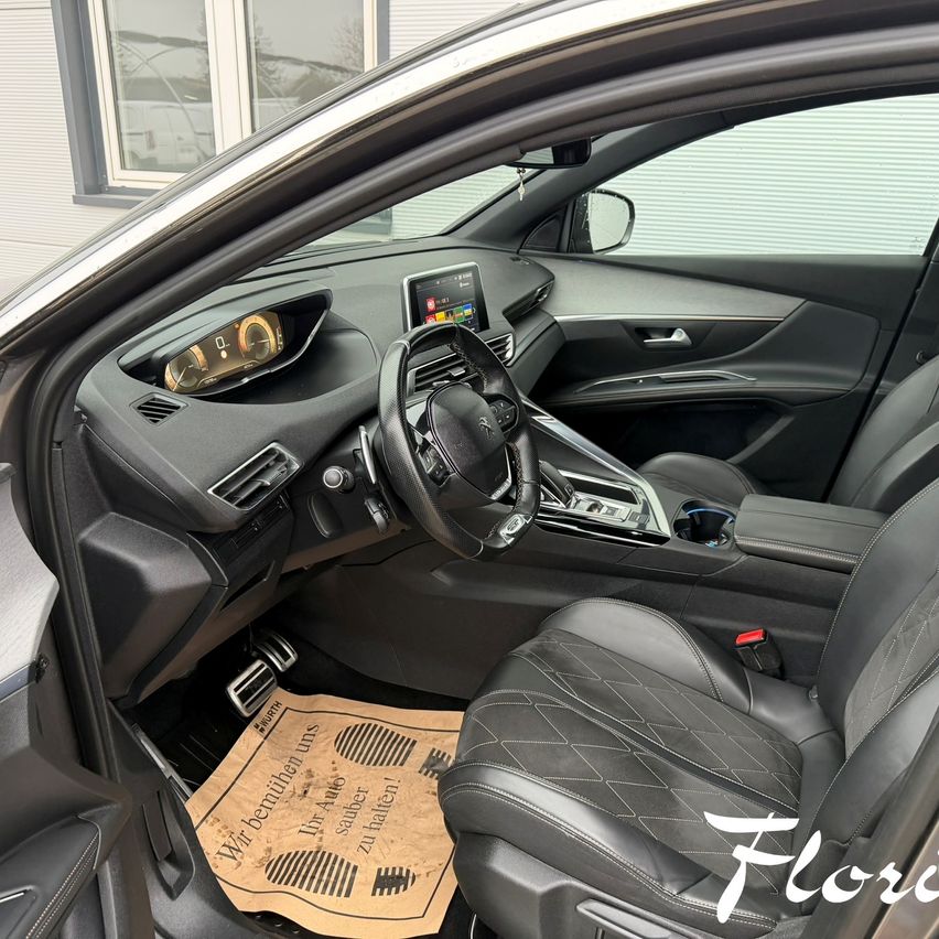 Das Innere eines modernen Autos ist sichtbar. Das Armaturenbrett zeigt einen Tachometer und andere Anzeigen. Das Lenkrad ist an Ort und Stelle. Ein Fußmatte zeigt 'Wir benutzen uns'. Das Auto ist vor einem Gebäude geparkt.