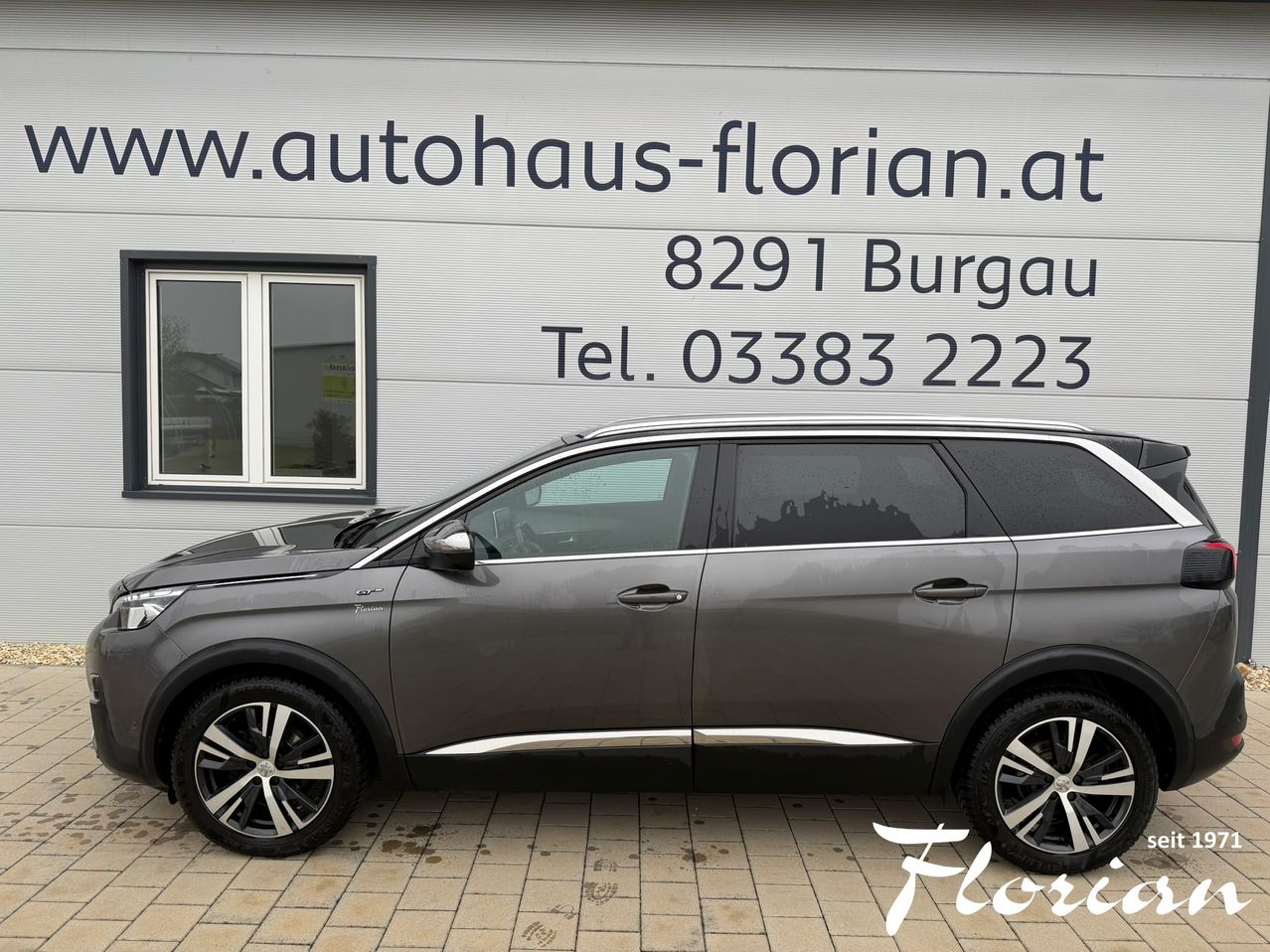 Ein grauer Peugeot SUV, der vor einem Gebäude geparkt ist, an dem ein Schild mit der Aufschrift 'www.autohaus-florian.at' und eine Telefonnummer '8291 Burgau Tel. 03383 2223' steht.