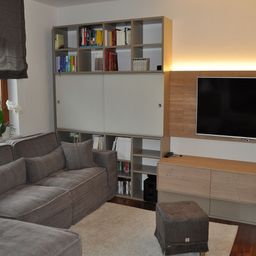 Ein modernes Wohnzimmer mit einer grauen Couch, einem Hocker, Teppich und einem hölzernen Entertainment-Schrank. Über der Couch befindet sich ein Bücherregal mit Büchern. Ein Flachbildfernseher ist an der Wand montiert.
